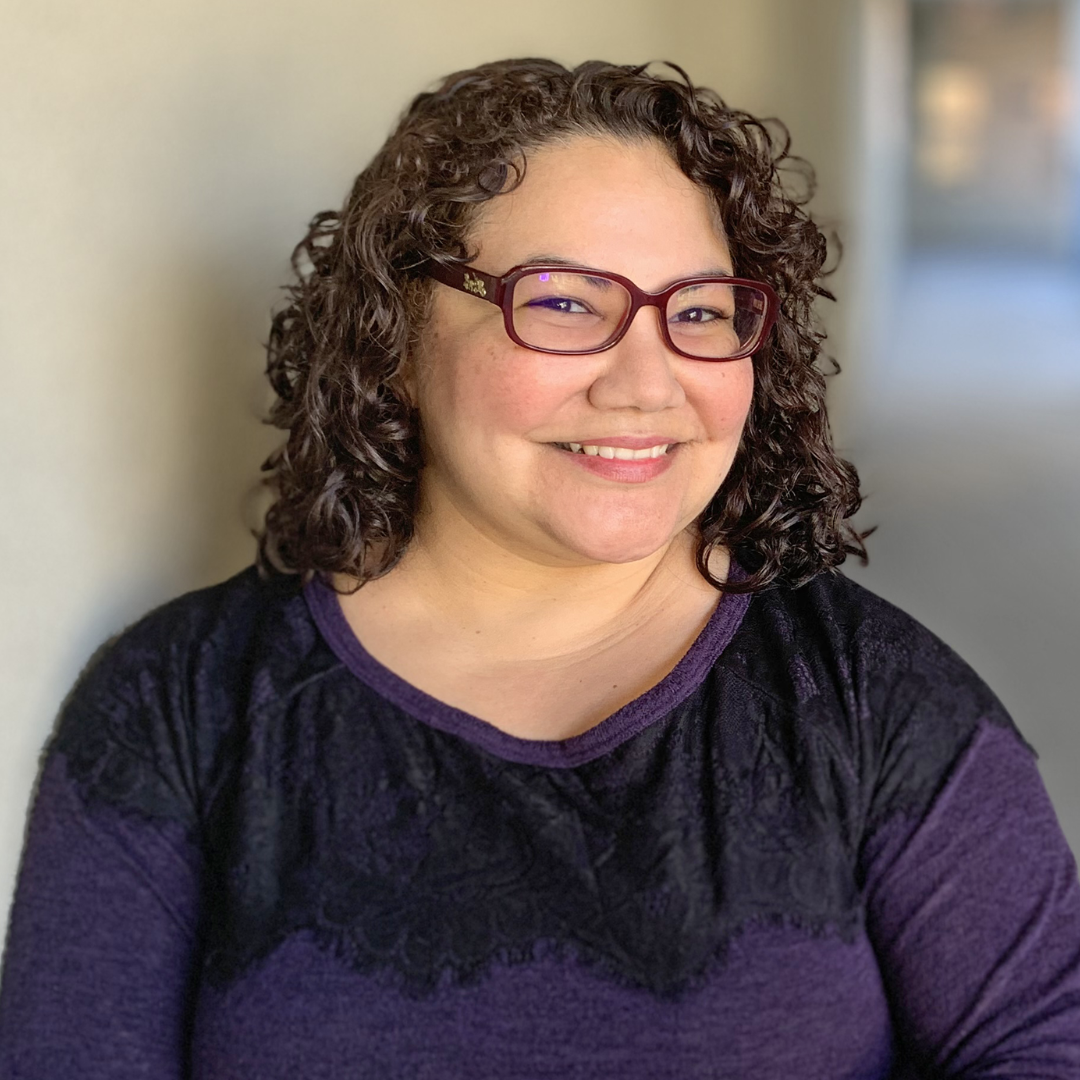 Paulette Erato, a woman with shoulder-length curly hair, smiles warmly at the camera. She is wearing dark-framed glasses and a purple top with black lace detailing. The background is softly blurred, with neutral tones that draw focus to her face.