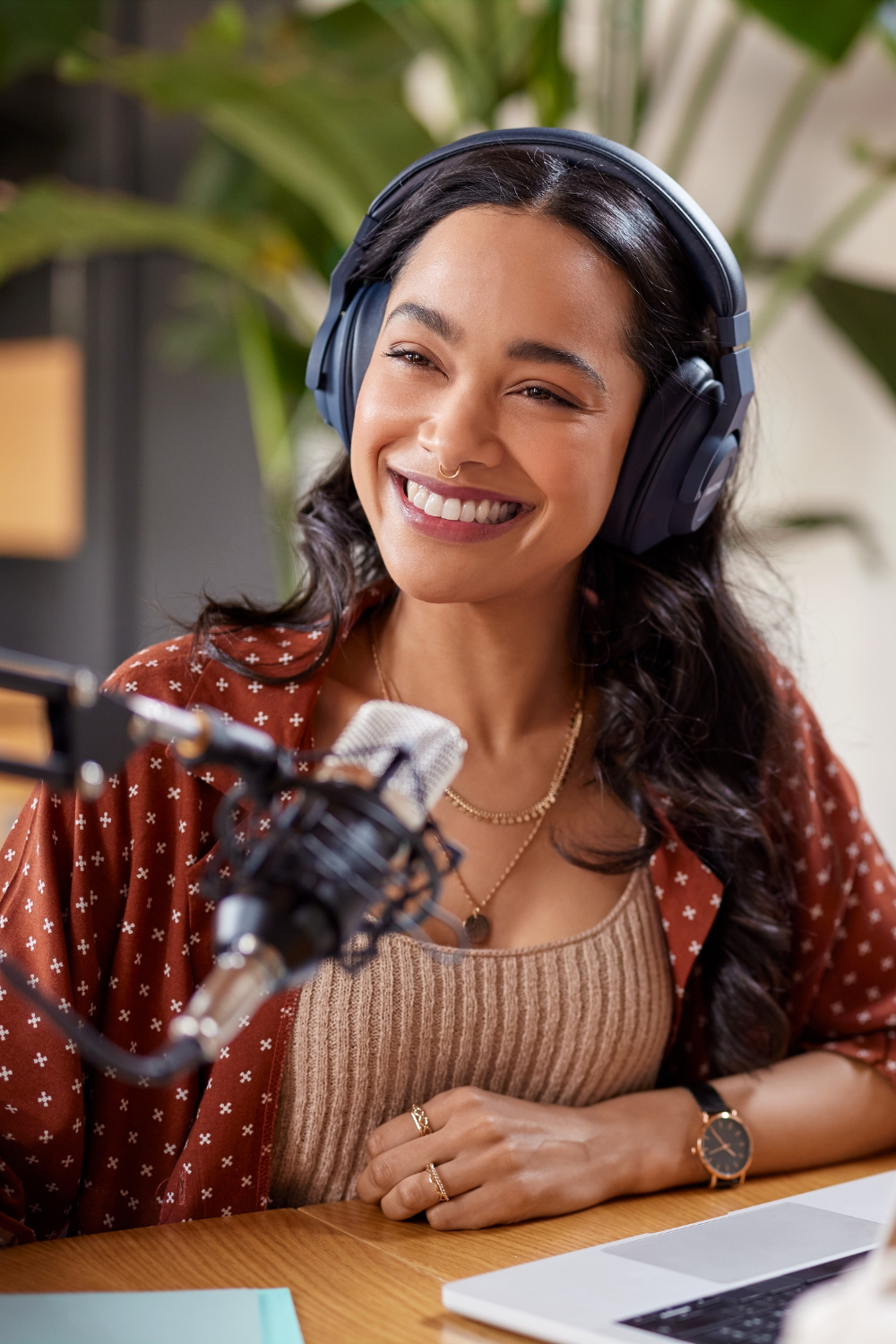 Brunette sporting a nose ring wearing headphones, a brown shirt with white stars and beige ribbed undershirt, and some gold necklaces smiles to someone off-camera. A microphone suspended by a mic arm is hovering in the air in front of her while a laptop sits open.