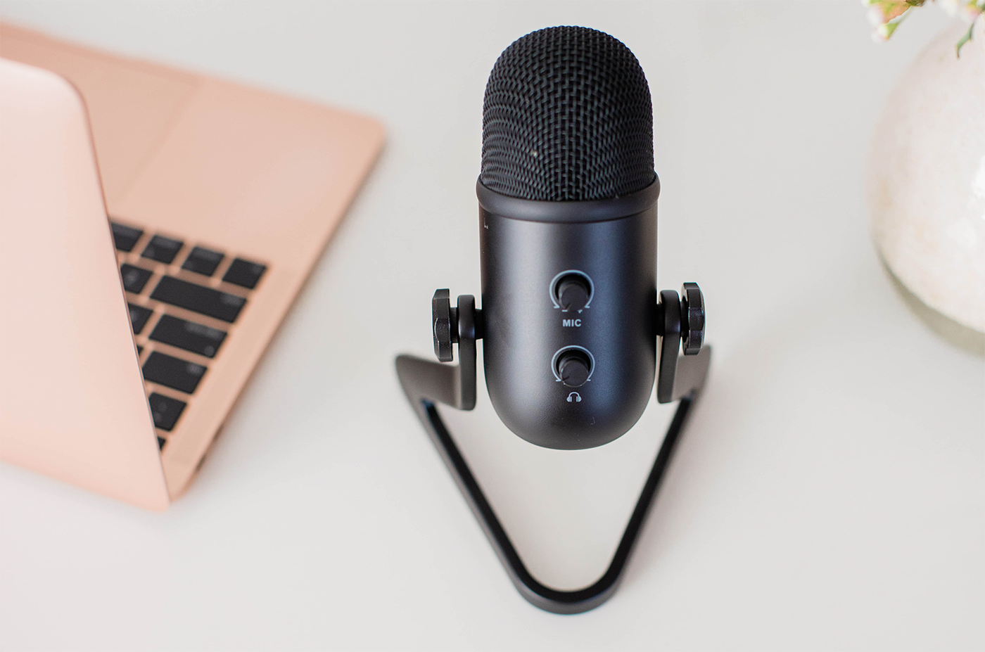 A minimalist podcasting setup with a close-up focus on a sleek black microphone placed on a white surface. The microphone features a sturdy stand and two control knobs for mic volume and headphone output. To the left of the mic is a partially visible pink laptop with a matte finish and black keys. In the background, there’s a soft blur of a vase with pale flowers.