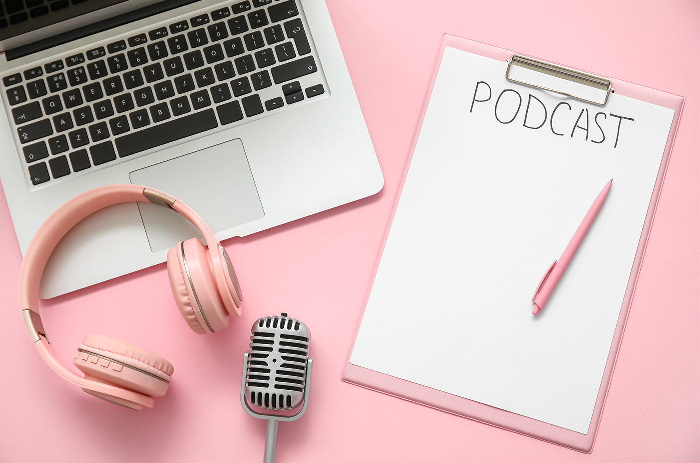 Flat lay of podcasting essentials set against a soft pink background. On the left is an open laptop with a silver body and black keyboard. To the left of the laptop is a pair of pink over-ear headphones, matching the color scheme. Below the headphones is a vintage-style silver microphone. To the right is a pink clipboard with a blank white sheet of paper that has the word "PODCAST" written at the top in bold black letters. A matching pink pen is placed diagonally on the clipboard.