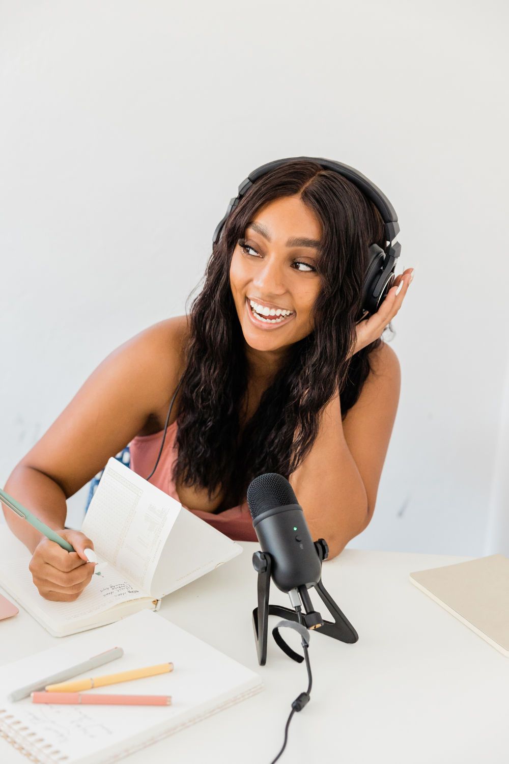 Young Black woman looking off-camera to the left while wearing headphones. She has one hand up to her ear while holding a pen and taking notes with her other hand. The microphone in front of her is on.