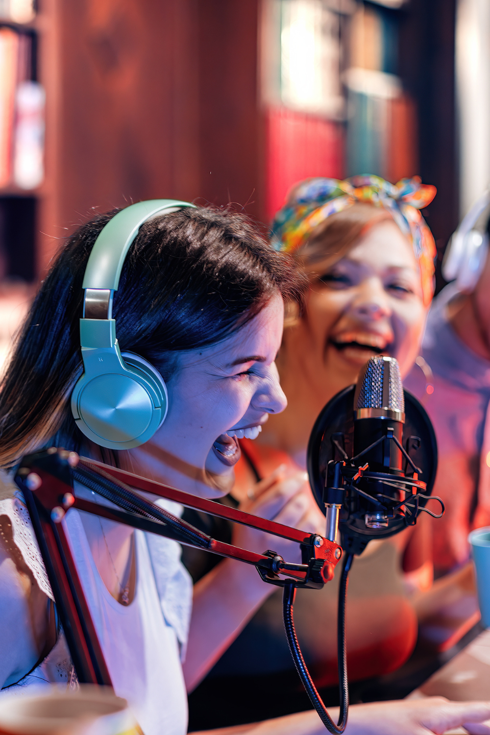 Two young women in frame are laughing joyously. The one nearest the foreground is wearing light green headphones and dark lipstick and is closest to the microphone being held by a mic arm. A little behind her the lady is slightly out of focus and wears a head wrap.