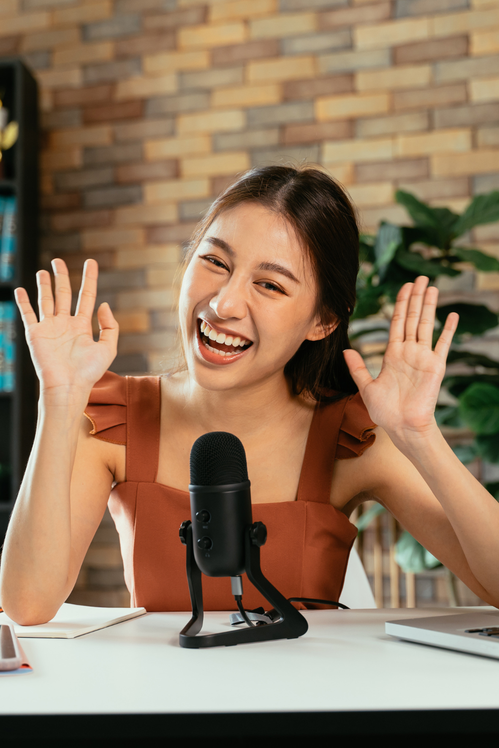 A young brunette smiles at the camera with a slight head tilt, her hands up as if to say hi! She has a black microphone on the white desk in front of her.